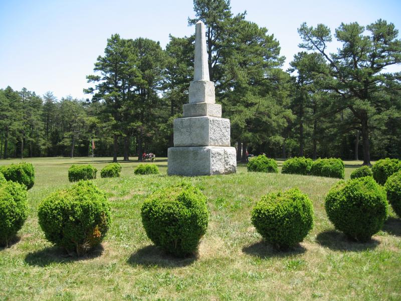 A small marble obelisk sits on three large blocks on a grassy hill. There are trees in the background and bushes in the foreground.