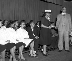 Charlotte Hawkins Brown on stage at Palmer institute with students.  There students seated behind her and she is shaking hands with a man in a suit. 