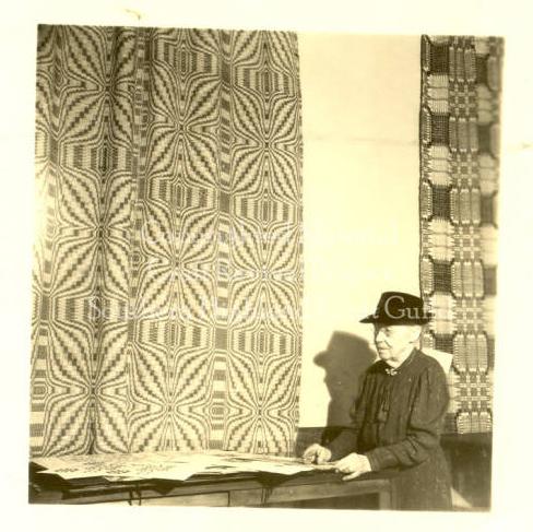 A woman sits at a table. A large tapestry with a psychedelic design hangs beside her. She is older and has glasses.