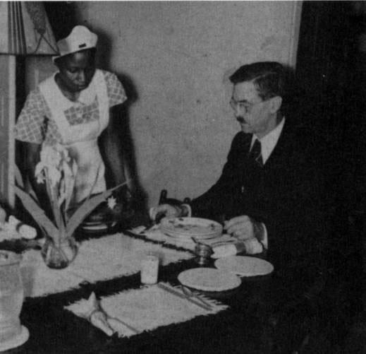 In this black and white photograph, a young black woman is serving Professor Caldwell at a dining table