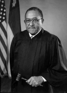Henry Frye. He is pictured wearing his judge gown and holding a gavel. He is straight-faced with glasses and short hair. 