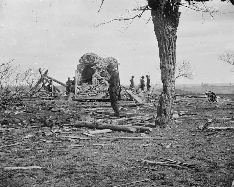 A ruined house in the middle of a battlefield. Fallen tree timbers litter the ground as soldiers survey the damage. Black and white. 