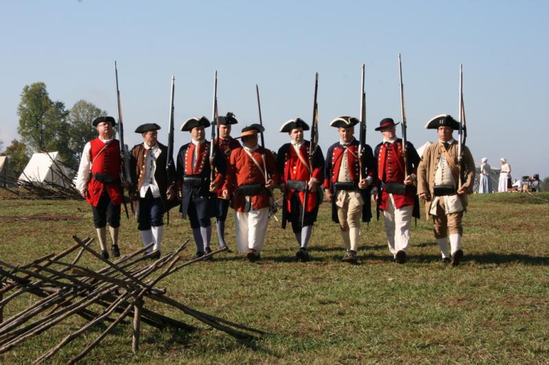 Provincial soldiers at Fort Dobbs Nine reenactor soldiers in a formation. They are wearing apparel of the colonial military, with tricorn hats and buttoned coats. It is sunny.