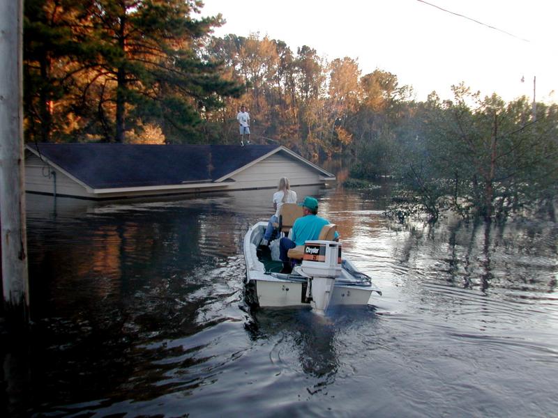 People ride a small boat through floodwater to rescue someone trapped on their rooftop. 