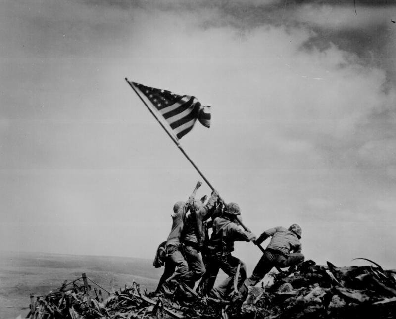 Marines raise a heavy flag on a rubble-covered hill. Black and white photo.