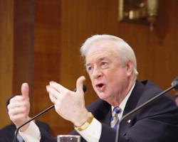 Jim Hunt speaking in a panel. He is older, wearing a watch with white hair and a suit. He speaking into a microphone.