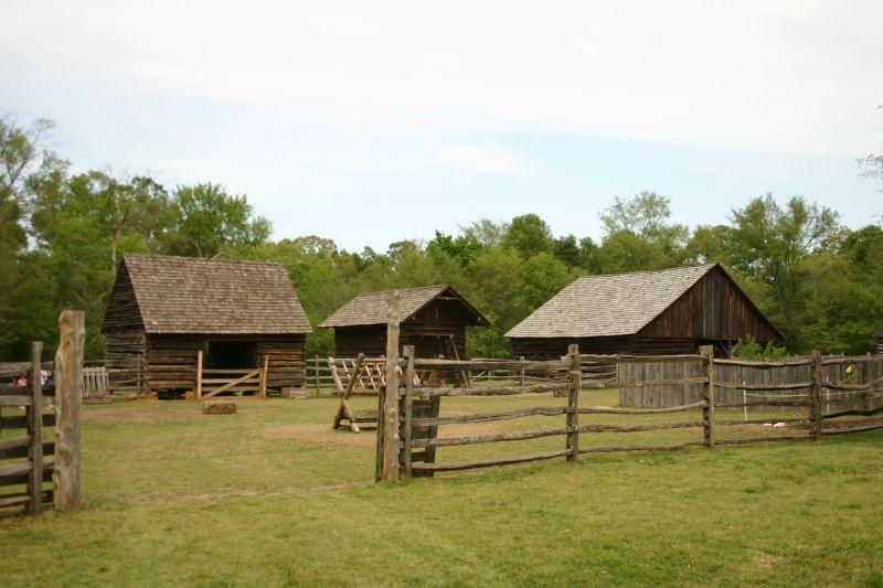 Latta Plantation outbuildings. There are three log buildings behind a timber fence. They are surrounded by woodland. There are clouds in the sky.