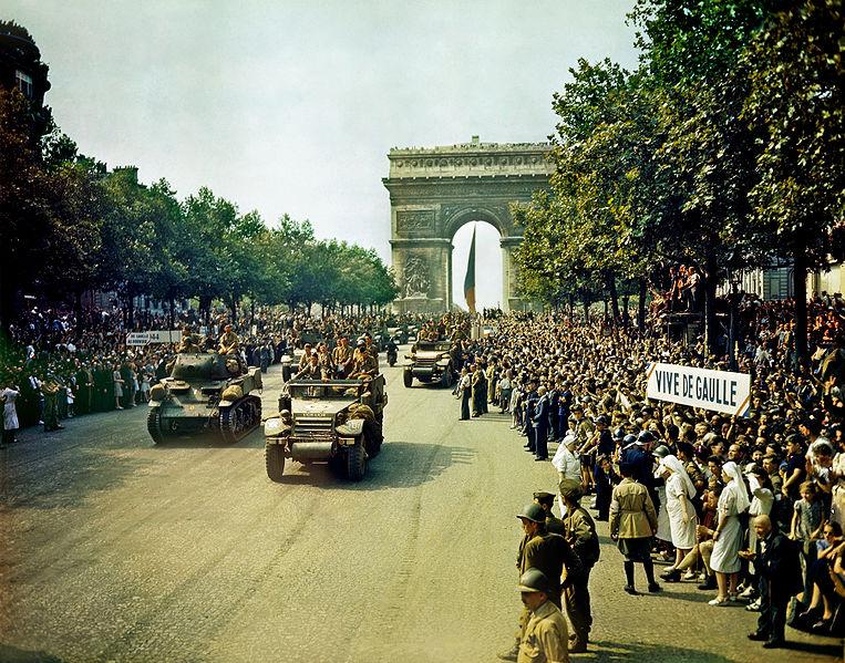 Soldiers marching down a crowded street of celebrators. The is a large stone Arch in the background. Signs with happy messages are held by the crowd. Color photo. It is sunny.