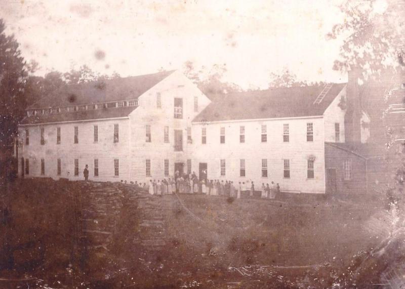 Alamance Cotton Mill Alamance Cotton Mill. It is a sepia print. There are people in front of the mill, which is a large building attached to a smaller building. There are trees around.