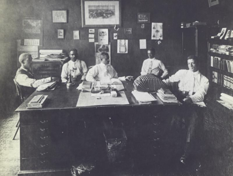 5 men in an office. There are pictures on the wall and office equipment on a large desk in the center of the room. The men are seated around the desk.