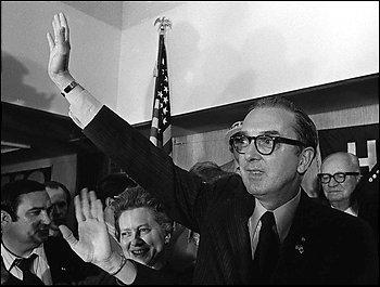 Jesse Helms celebrating his victory. He is pictured waving to a crowd. He has glasses, a suit, slicked hair, and no facial hair. He has a triumphant expression. 