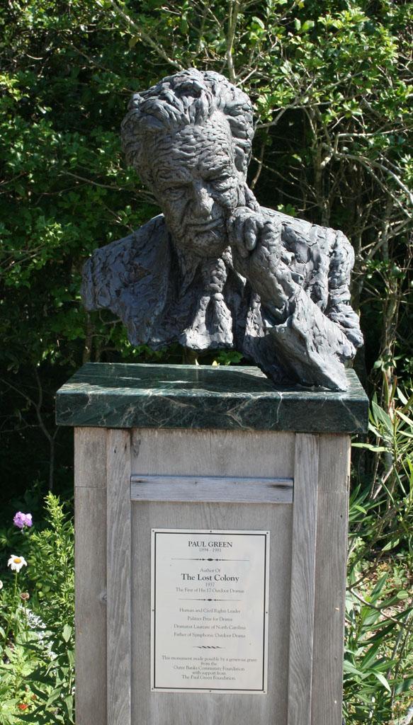 Metal statue of Green. His finger is drawn and pressed to his face. Trees are behind the bust. It is a sunny day. 