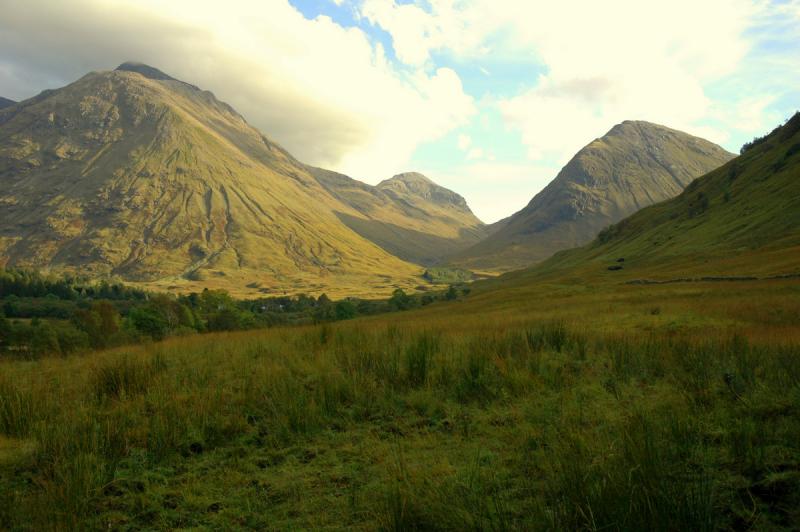 Scottish Highlands. Rolling green hills with mountains in the background. The sky is cloudy but blue.