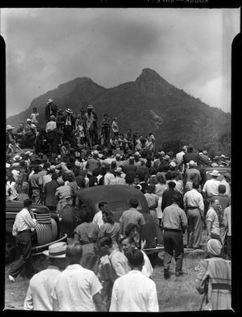 People gather in front of a mountain together. There are a few cars in the middle. Black and white. 