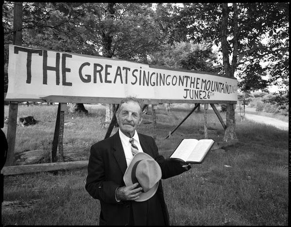 Black and white photograph of Joe Hartley, holding his hat in one hand and a book in the other, in front of a handwritten sign that says "THE GREAT SINGIN ON THE MOUNTAIN JUNE 26"