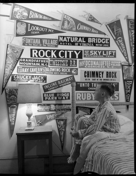 This black and white photograph is of Hugh Morton's son, Jim. He is sitting on his bed in his pajamas with his arms clasped around his knees as he looks at the tourism stickers he has collected on his wall.