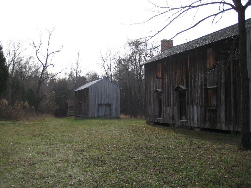 An old, brown wooden house beside another small gray wooden house. They are adjacent to a forest where none of the trees have their leaves. The sky is misty. 