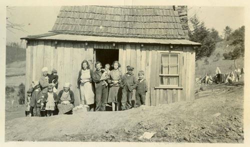A large family stands in front of a small shack. 
