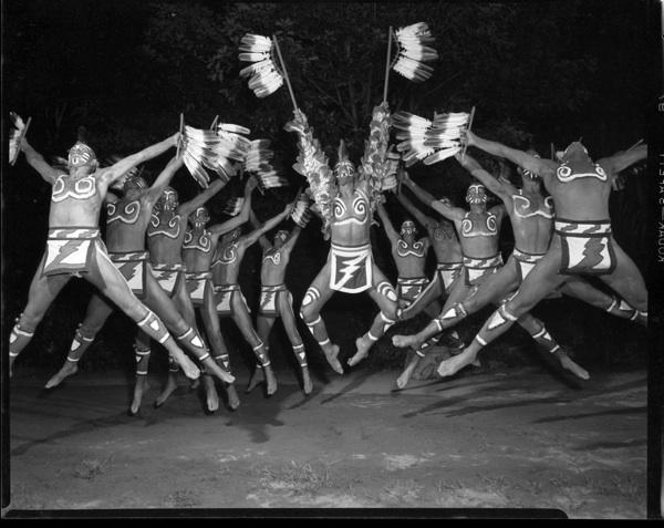 Men costumed in feathers and in loincloths are caught suspended above the ground, in mid-jump.