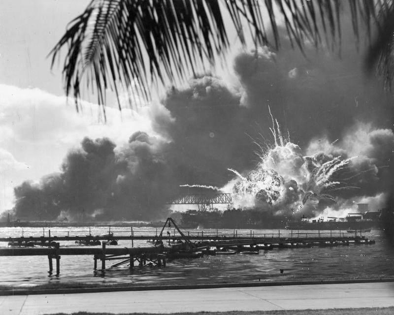 black and white photo of a battleship exploding in a harbor. A palm tree is in the foreground. 