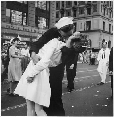 A sailor kisses a nurse on a busy street. They are both wearing their work apparel.