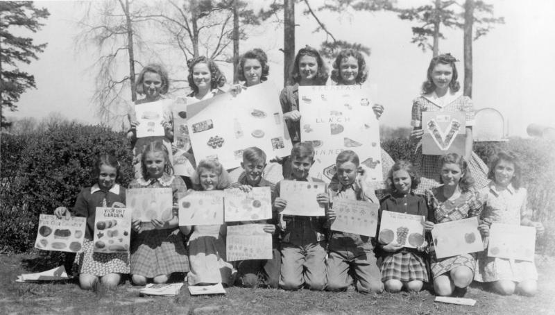 A group of 15 students are pictured in this black and white photograph showing their 4-H posters which celebrate “Victory Gardens.” Six girls are standing behind a group of kneeling boys and girls in front of some tall pine trees and a hedge.