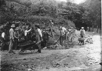 Photo. Men work to repair a road. Their are carts, tools, and other equipment about. The hillside they are working near is muddy.
