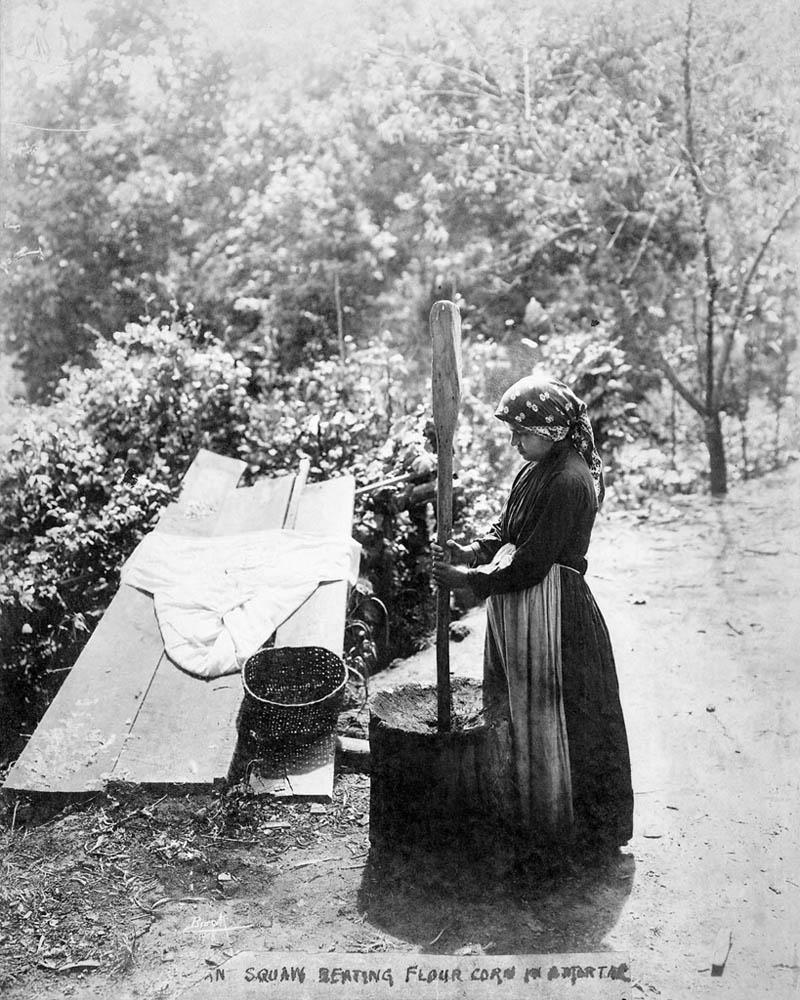 Cherokee Woman Grinding Corn in a Mortar Black and white photograph of an American Indian woman pounding corn with a large mortar and pestle.
