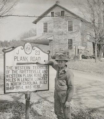Mr. John A. Shore holding a piece of the plank road in front of the North Carolina Highway Historical Marker in Bethania, 1959. Mr. Shore owned the general store in the background.