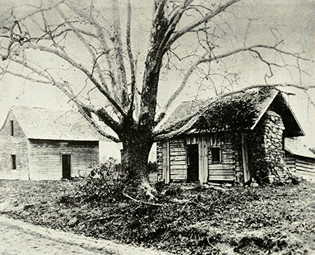 A small log shack beside a large tree with a medium shack in the background. This is Bennett Place. Photo.