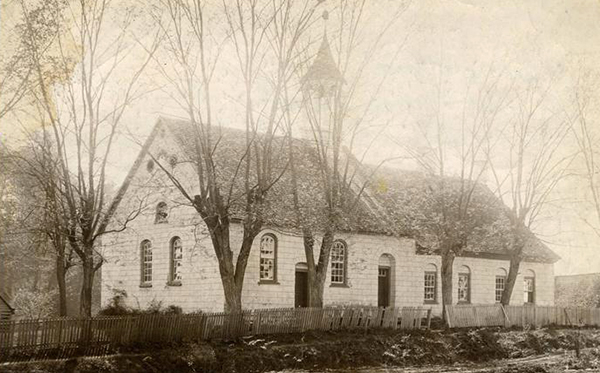 A sepia photo of an old church in a woodland. It is white with a black roof.
