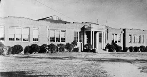 A long one-story brick building.  There is a columned entryway in the middle of the building and many windows to each side of the entry.  There are short bushes under the windows.