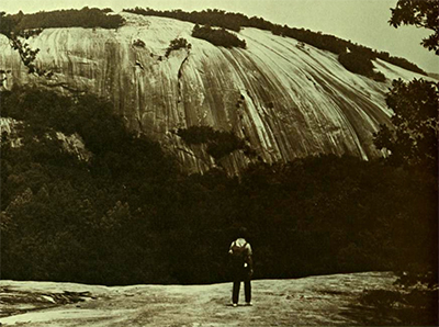 Hiker looking up at rock face. The rock face has brush growing throughout.