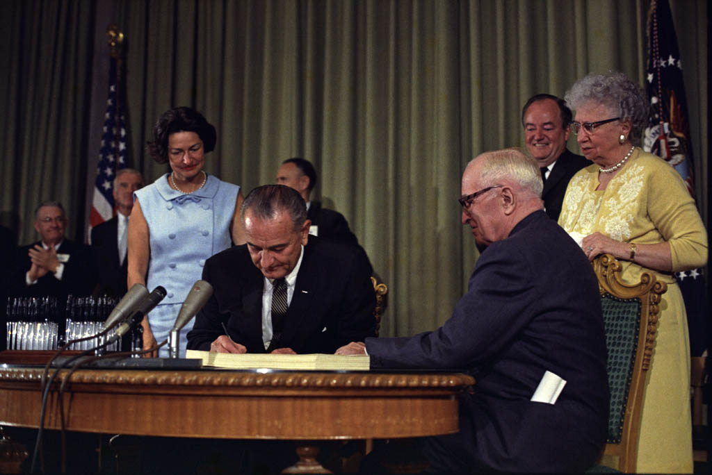 President Johnson signs legislation at his desk. Other activists of the bill surround him.