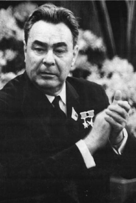 Leonid Breznev. He has medium hair and a stern expression. His hands are clasped and he is wearing a suit.