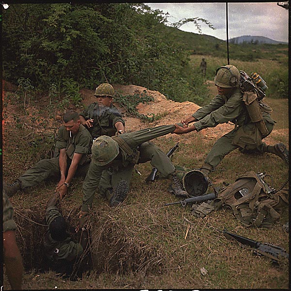 Two soldier lower a third into a bunker. They are all wearing tactical equipment.