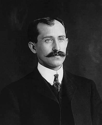 Orville Wright. He has a handlebar moustache and is wearing a suit. He has coifed, short hair.