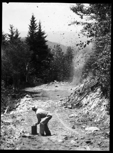 A road being blasted from rock with dynamite on a mountainside. Black and white photo.
