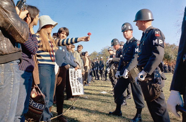 Protestors stand in line with military police while protesting. They all stand on a sunny grass lawn. They military police are heavily armed.