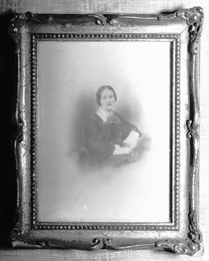 A woman in a dark colored dress is sitting looking toward the viewer. She is wearing a dark dress with white at the collar and the wrists. Her dark hair is parted in the middle and pulled back and secured at her nape.