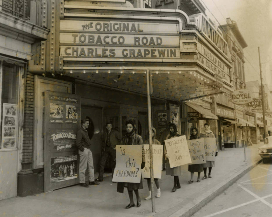 Five young women holding protest signs picket outside of the National Theatre. White men bystanders laugh in the background.
