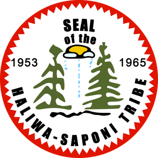 A badge. It reads "Seal of the Haliwa-Saponi Tribe, 1953/1965." Two pine trees are separated by a raining cloud with a sun behind it. The badge is bordered by a jagged, red edge.