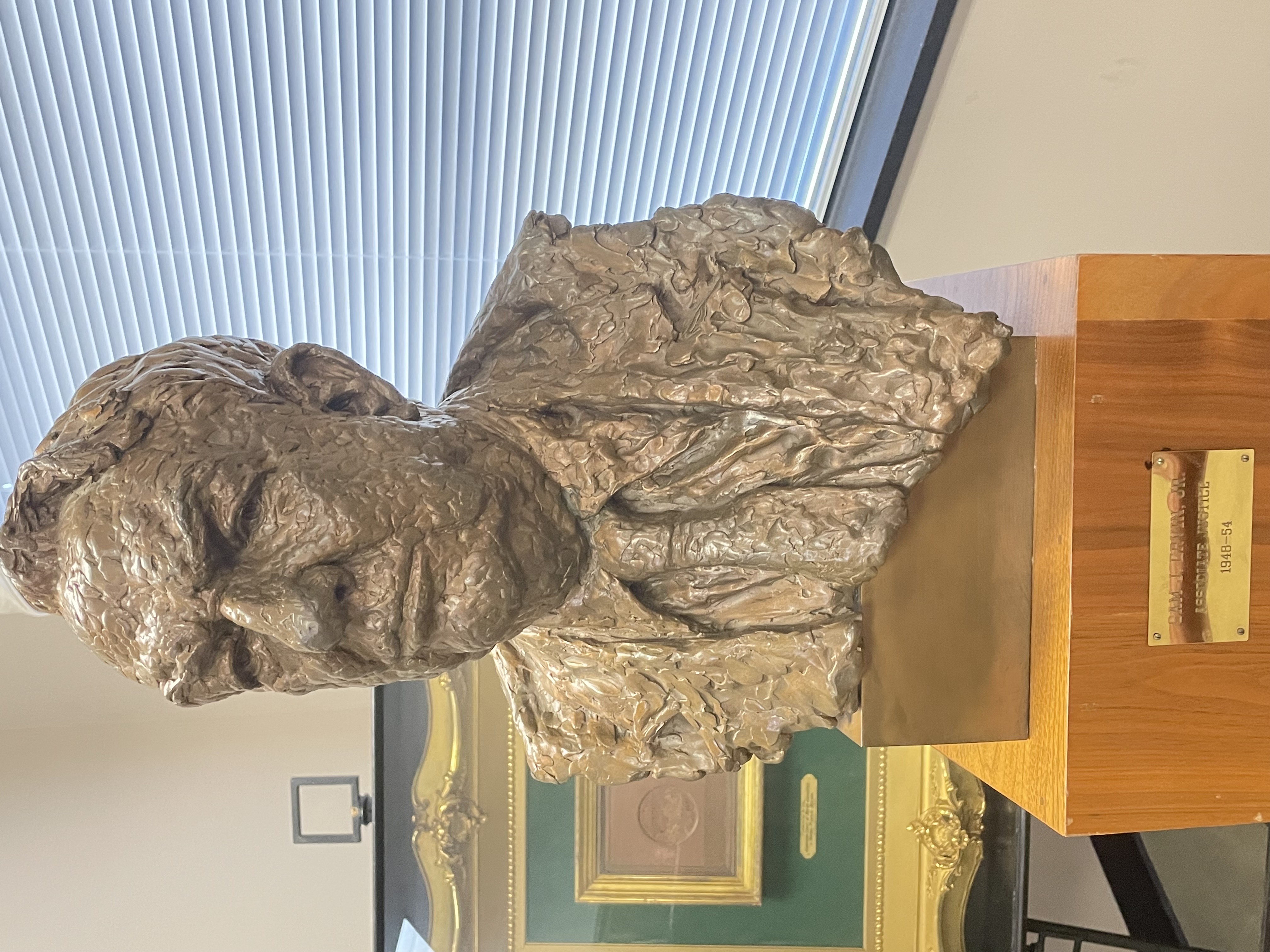 A metallic bust sits on a plinth. It depicts a man wearing a suit and tie.