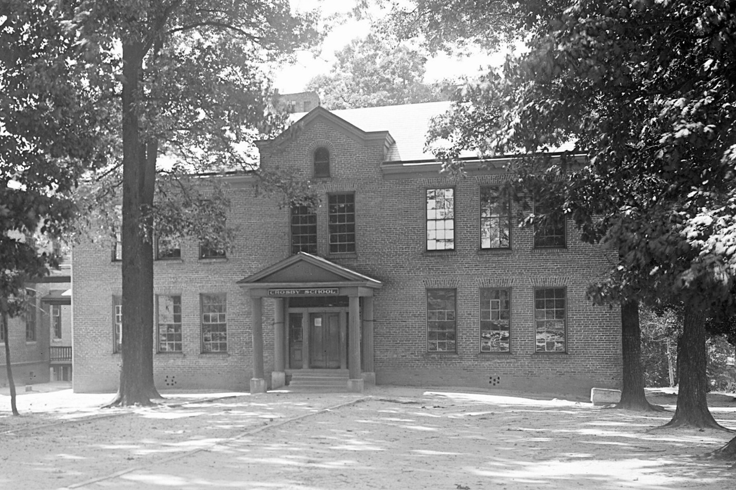 A brick building with 14 windows. Above the door, a sign reads 'Crosby School." Large trees flank all sides of the building.