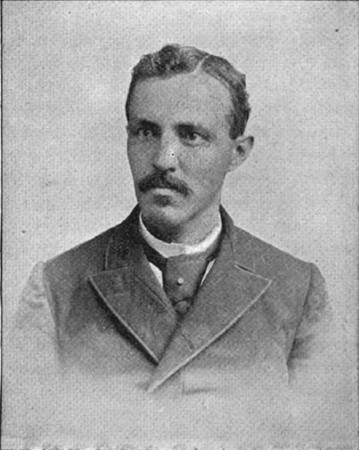 Black and white photograph of John Crosby. He has a mustache and small chin beard, his hair parted on the right side.  He is gazing to the left with a serious expression. He is wearing a fastened jacket, white shirt and tie.