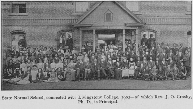 Students and staff (about 75) pose for a photo in front of a brick building. They are wearing formal wear like dresses and suits.