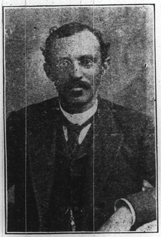 Crosby sits facing the camera.  He has a mustache and soul patch.  His hair is dark and parted on the right side. He is wearing round wire-rimmed glasses. He is dressed in a suit coat, vest, white button-up shirt and a tie.