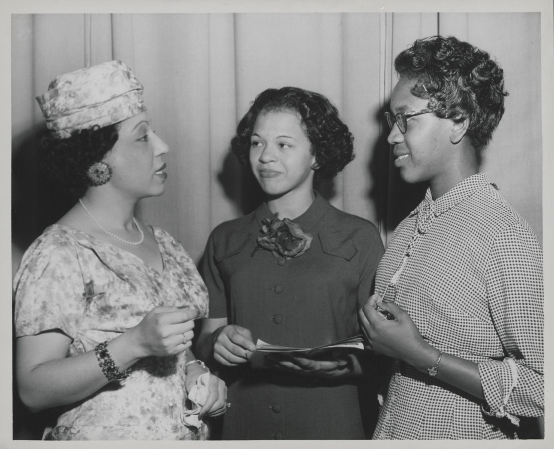 Elreta Alexander talking to two other women. She is pictured far left, appearing to be mid sentence. All women are dressed in nice clothes.