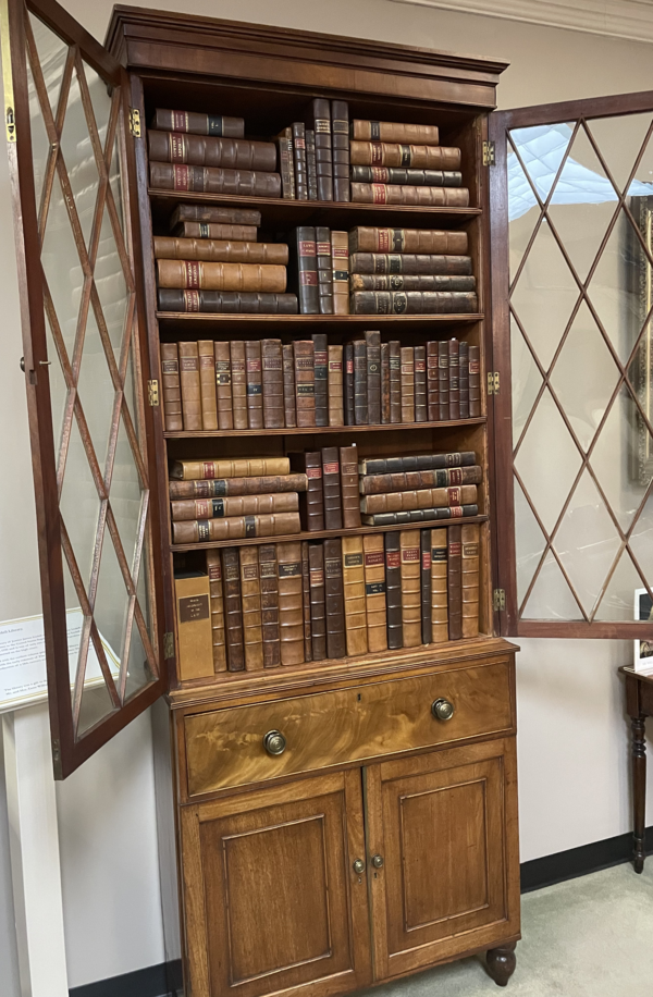 A bookcase with leather books stuffed all throughout. Its glass cabinet doors are opened.