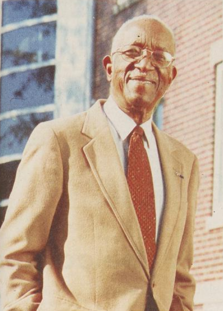 John Hope Franklin wearing a tan suit and smiling. He is older, and a brick building is behind him.